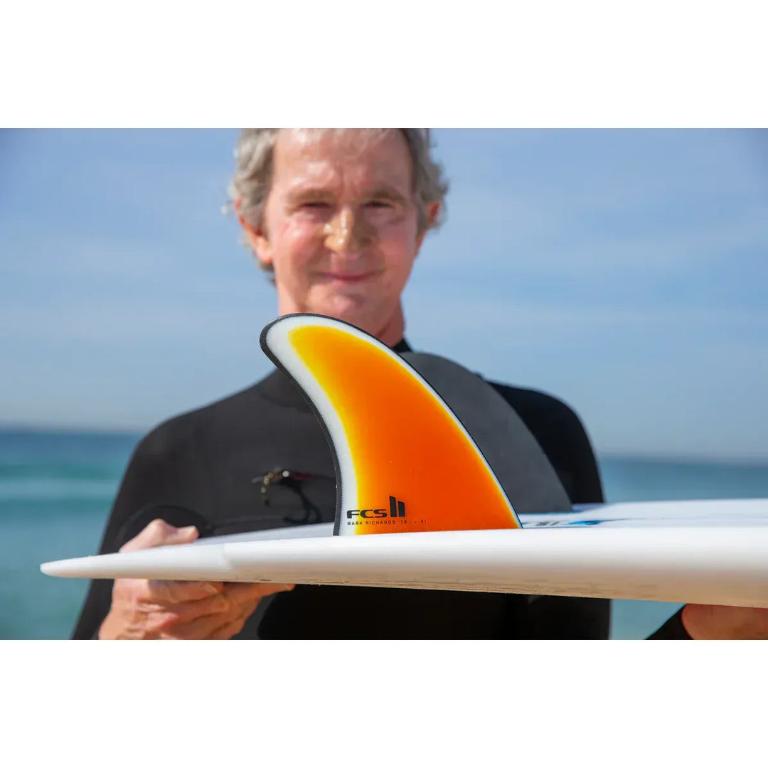 Man holding a surfboard with a visible FCS logo on an orange fin against a clear sky.