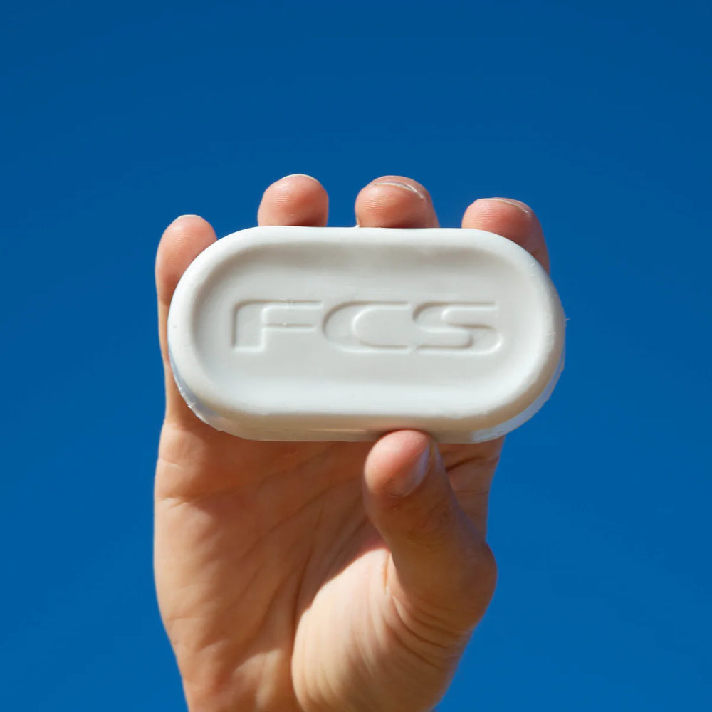 Hand holding a bar of surf wax with 'FCS' branding against a blue background