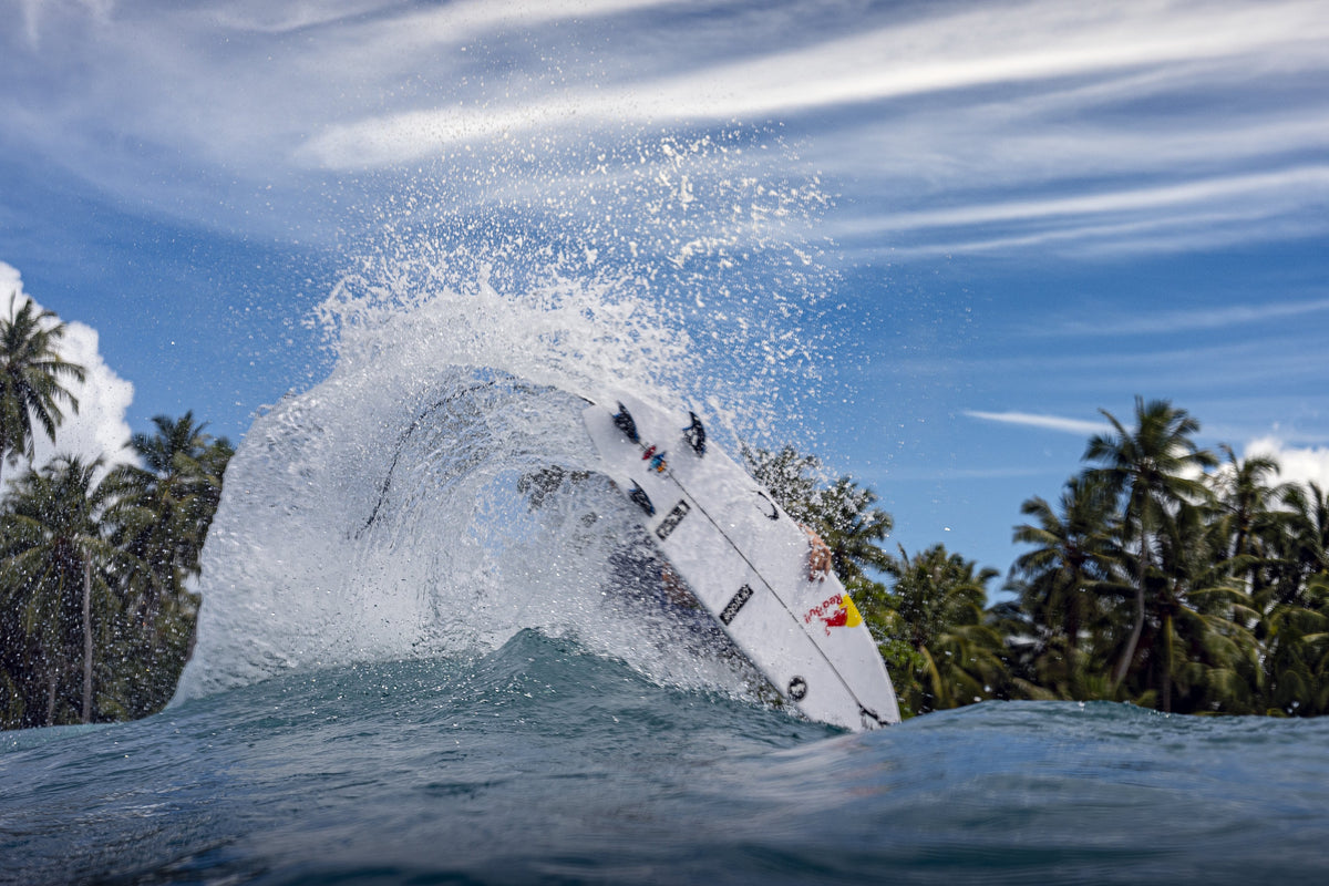 Photograph from the rear of a wave showing the bottom of a surfboard, showing several sponsor logos and fins above the wave, with a plume of spray extending behind the board.
