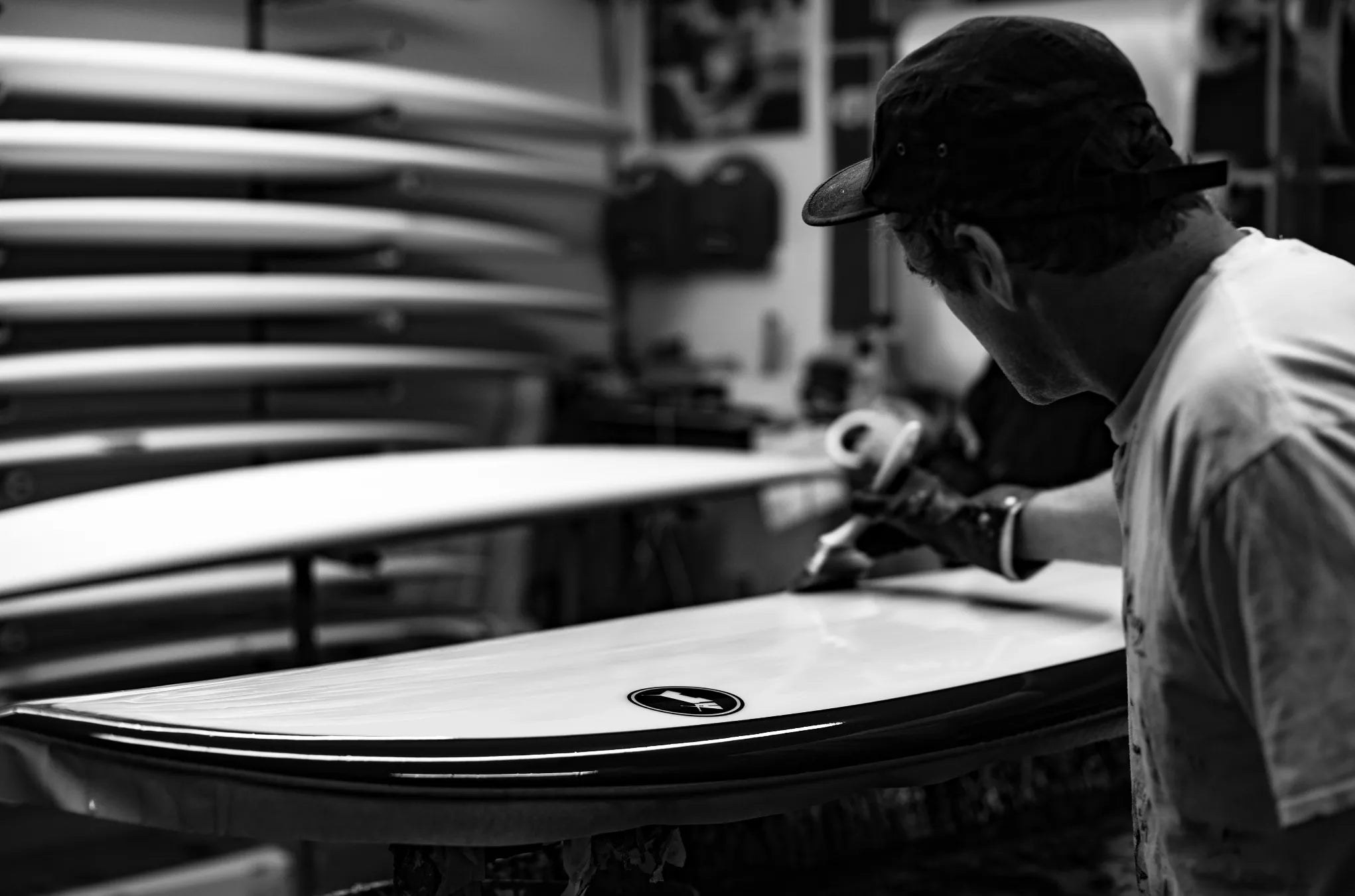 Surfboard laminator working on a surfboard in a workshop with stacked boards in the background