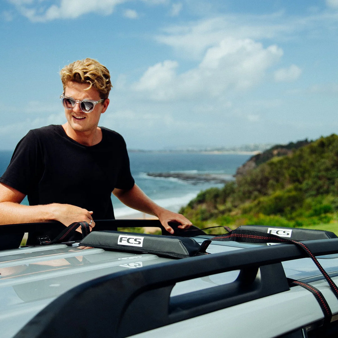 Man standing next to a car with FCS surfboard racks on a scenic coastal road.